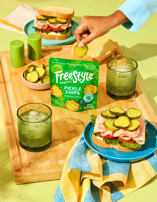 Table setting with sandwiches, pickles, and Freestyle Snacks Pickle Chips on a wooden board.