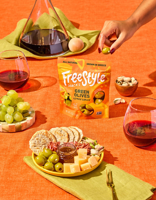 Table setting with Freestyle Green Olives Lemon Garlic, wine, and snacks on an orange tablecloth.
