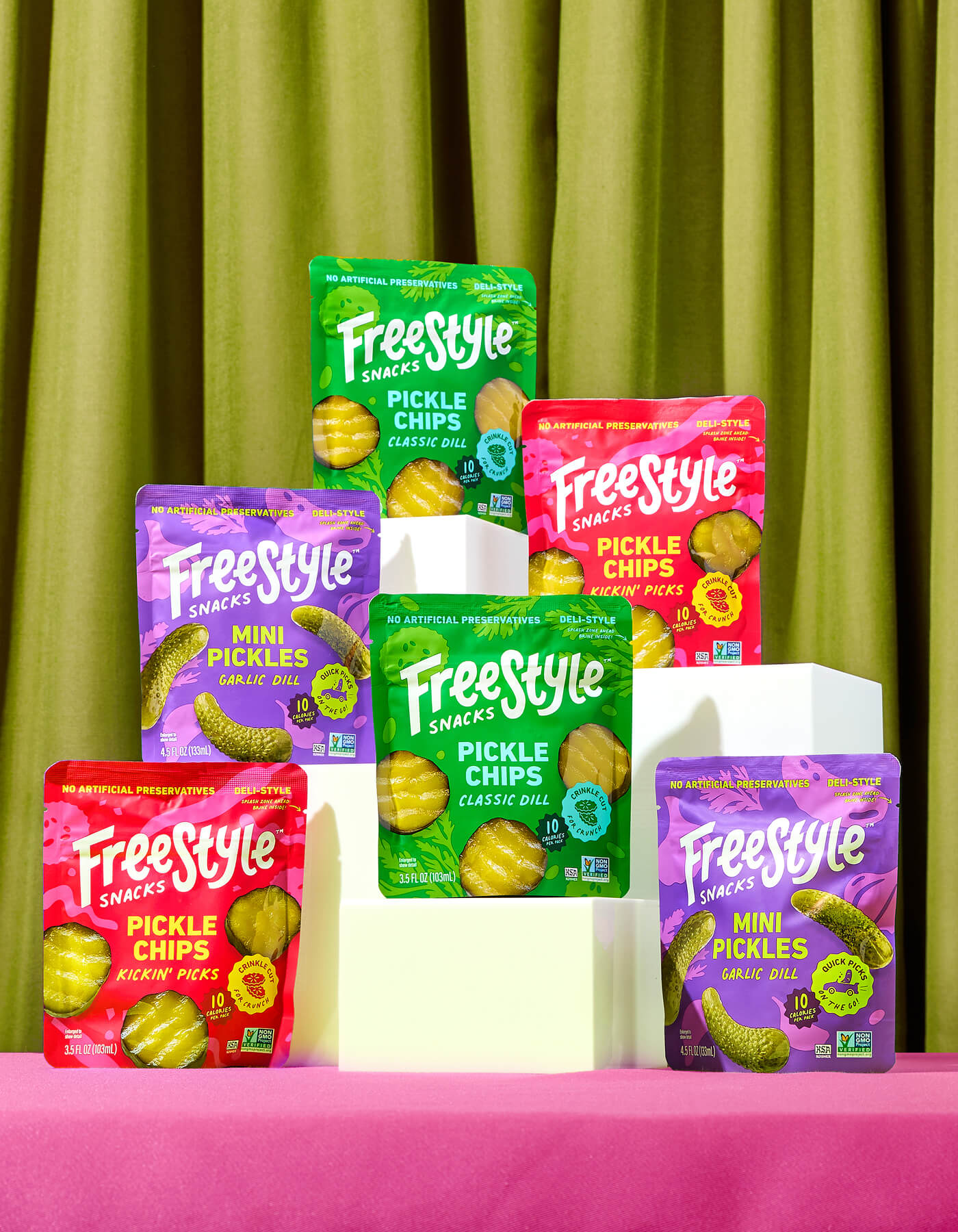 Assorted Freestyle Snack Pickle Packages on a pink surface with a green curtain background – 2 Packs of Mini Pickles Garlic Dill, 2 Packs of Pickle Chips Classic Dill, 2 Packs of Pickle Chips Kickin' Picks